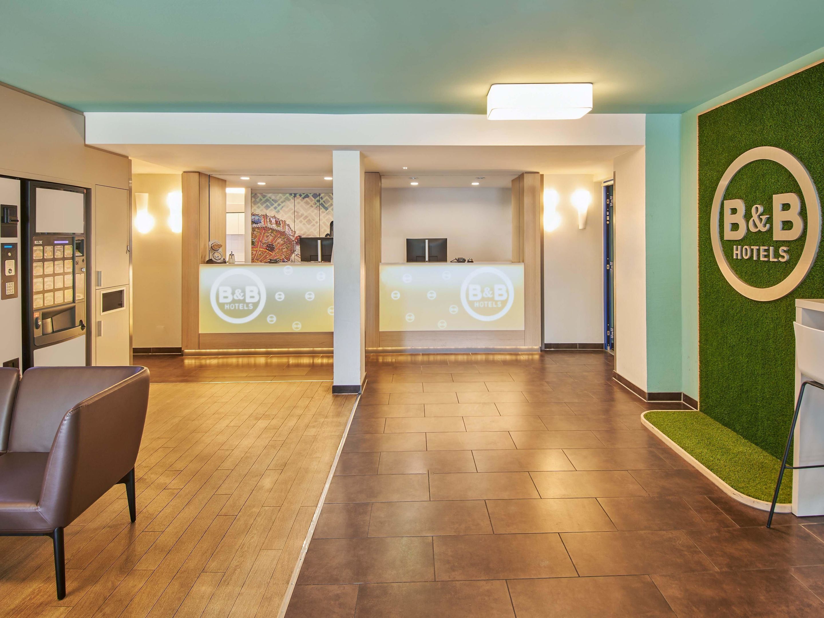 Zona de recepción con el letrero de B&B Hotels, mostrador de recepción, área de espera con una silla y una pared verde con césped artificial.