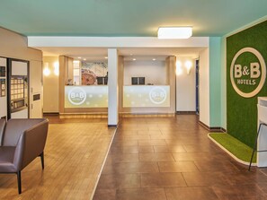 A reception area with a B&B Hotels sign, a reception desk, a waiting area with a chair, and a green wall with artificial grass. - B&B Hotel Darmstadt (Darmstadt)