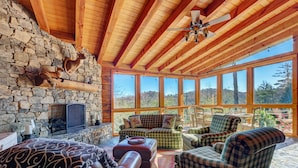 A cozy cabin interior with a stone wall, wooden ceiling beams, a fireplace, and large windows offering a view of the forest. - Breathtaking views,private property,glass porch w/wood-burning fireplace (Highlands)