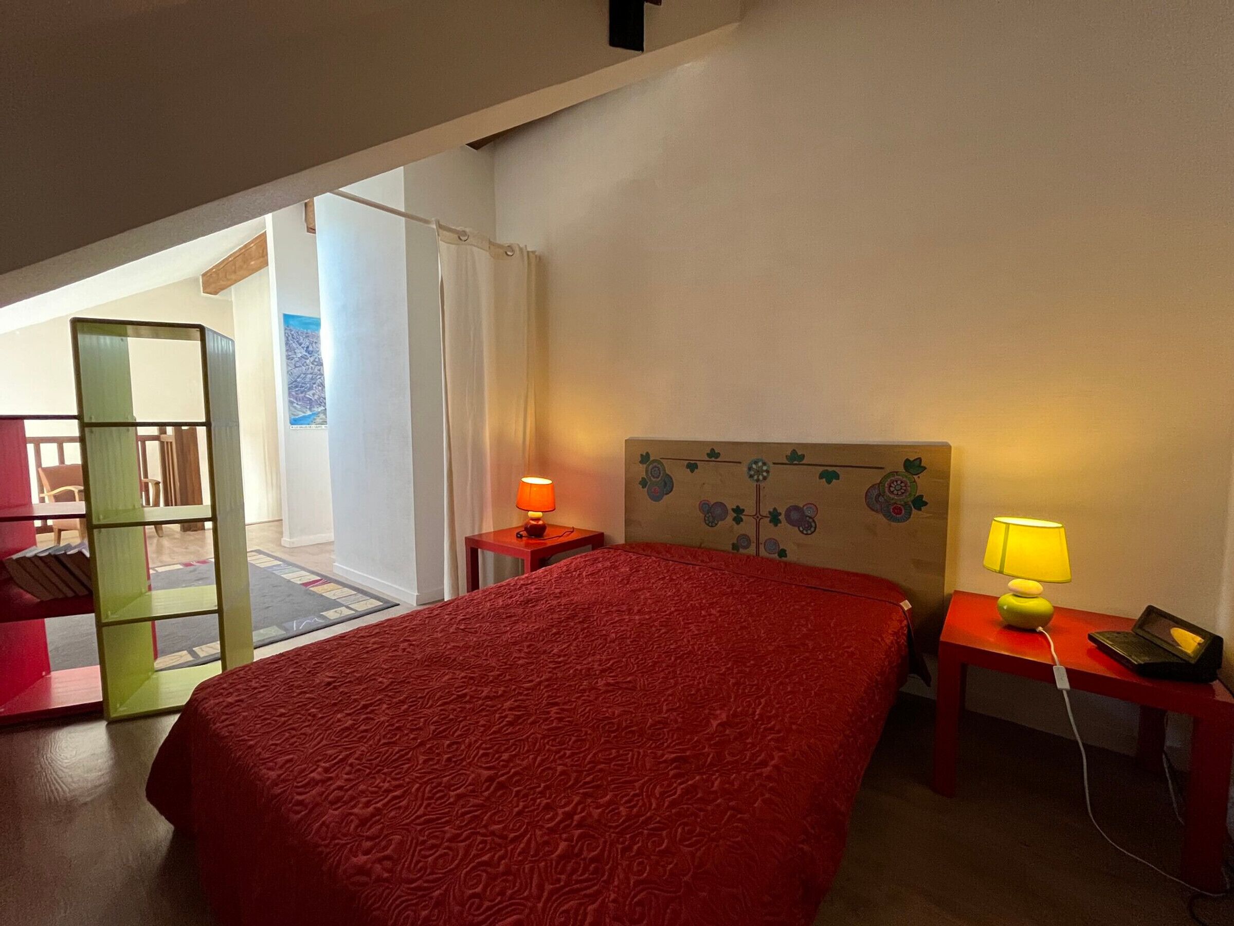 Ein Hotelzimmer mit rotem Überwurf, zwei Nachttischlampen, einem hölzernen Kopfende mit floralen Verzierungen, einem kleinen Tisch mit Lampe und Blick auf einen Balkon mit Stuhl und Tisch.