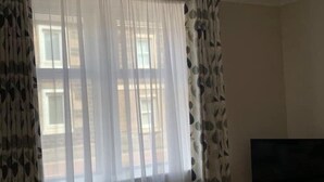 Et værelse med et stort vindue med lette gardiner, et fladskærms-tv på et stativ, og en mønstret gardin til venstre.