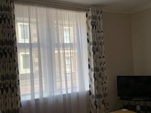 A room with a large window covered by sheer curtains, a flat-screen TV on a stand, and a patterned curtain on the left side. - No 67 Leuchars (Nr St Andrews) Free Parking - Pet Friendly (Leuchars)