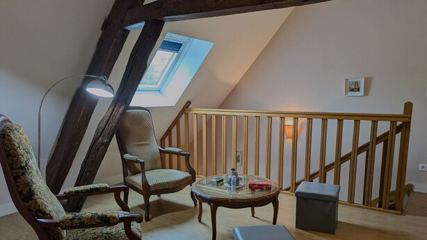 Une chambre à coucher confortable, aménagée en mezzanine, avec un plafond en pente, des poutres en bois, une petite table ronde avec une lampe, deux chaises et un banc.