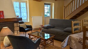Un salon chaleureux avec un canapé, un fauteuil et une table basse. On aperçoit un escalier au fond.