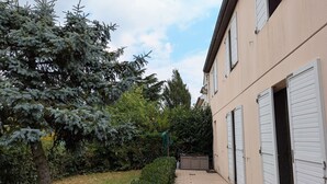 Un estrecho sendero pavimentado que conduce a un edificio con contraventanas blancas, rodeado de follaje y un gran árbol de hoja perenne.