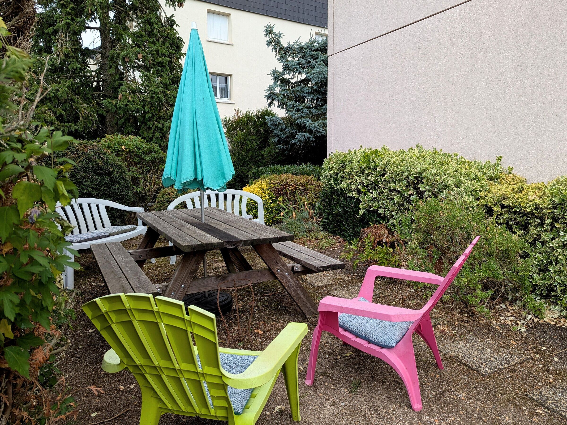 A wooden picnic table with a turquoise umbrella, surrounded by colorful plastic chairs.