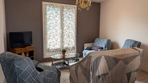 Una sala de estar con televisor, dos sillones estampados, un sofá, una mesa de centro de vidrio y una araña.