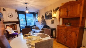 Een gezellige woonkamer met een open haard, een eettafel gedekt voor twee personen en uitzicht op het landschap door grote ramen.