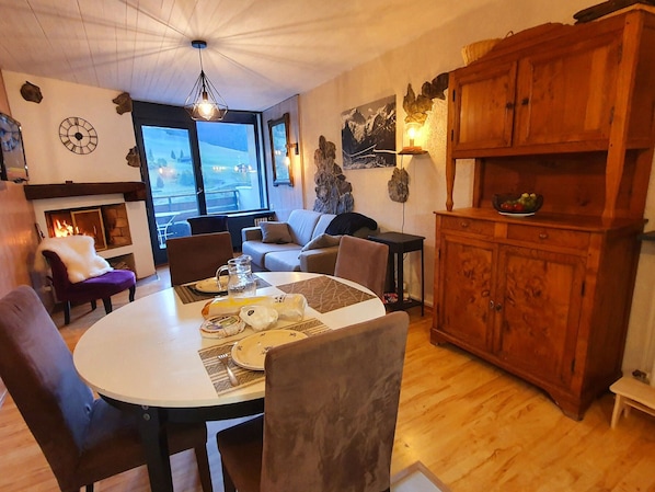 Un salon chaleureux avec une cheminée, une table à manger dressée pour deux personnes et une vue sur l’extérieur grâce à de grandes fenêtres.