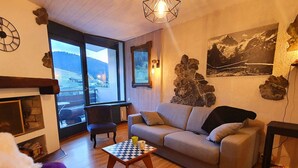 Een gezellige woonkamer met een bank, een kleintje tafeltje en een open haard. Er is een groot raam met uitzicht op bergen en een auto geparkeerd buiten.