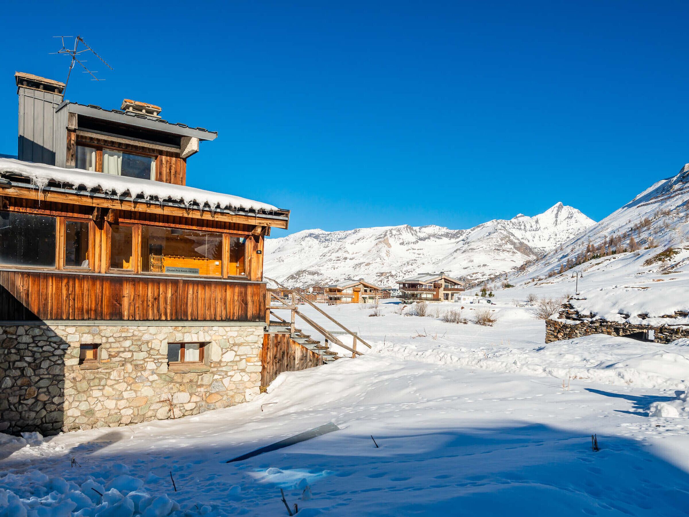 A wooden chalet with stone walls, a ski lift, and snow-covered mountains in the background.