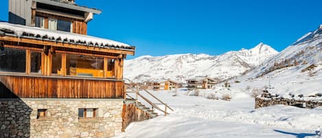 A wooden chalet with stone walls, a ski lift, and snow-covered mountains in the background.