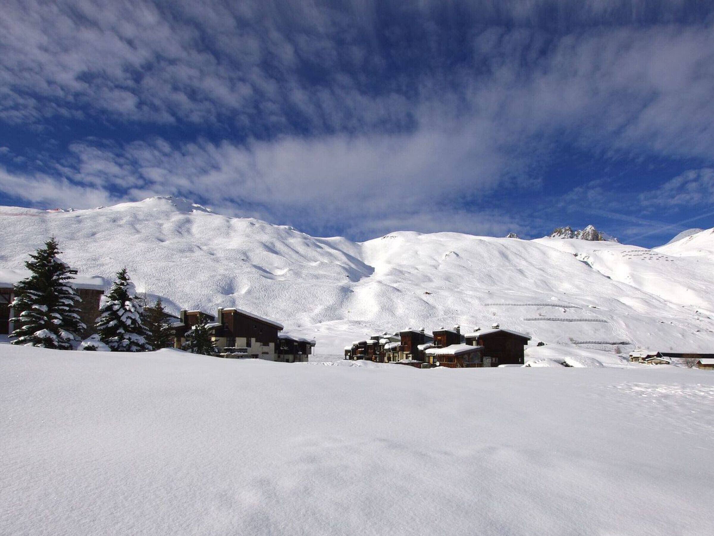 Una montagna innevata con cielo sereno. In primo piano si vedono edifici dalle tegole imbiancate dalla neve e alberi.