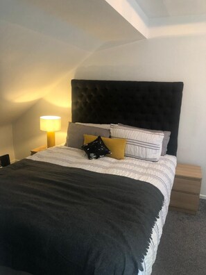 A neatly made bed with a dark headboard, white and striped bedding, and a black throw blanket. There is a bedside lamp on the left and a wooden nightstand with drawers. - The 5 bed House With Spacious Rooms and Lounge (Coventry)