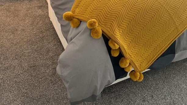 Un lit bien fait, aux couleurs gris et jaune, une table de chevet en bois avec une bougie, et un mur sobre.