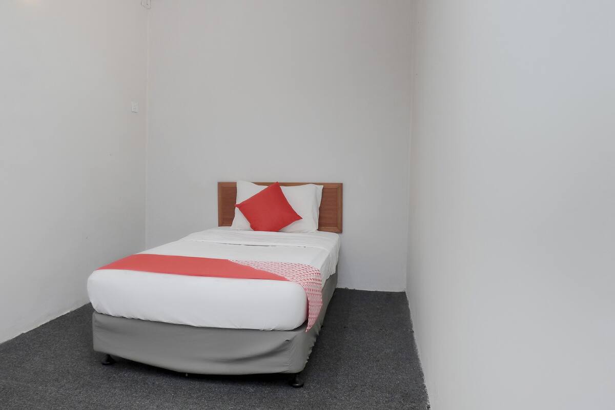 Een kleine kamer met een enkel bed, een houten hoofdbord, wit beddengoed en een rood kussen.
