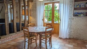 A rustic dining area with a wooden table and chairs, a brick fireplace, and a large window with curtains. - Charming 2-Bedroom Rural House with Private Terrace Near Fontainebleau (Lorcy)
