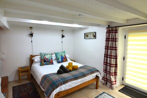 A bedroom with a wooden bed, a small bedside table, a wall-mounted lamp, a patterned rug, and a window with curtains.