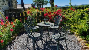 Een terras in de tuin met een ronde tafel en vier stoelen, omringd door diverse bloeiende planten en een stenen muur.