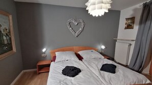 A double bed with white bedding, two bedside tables with lamps, a heart-shaped wall decoration, a radiator, and a framed picture on the wall.