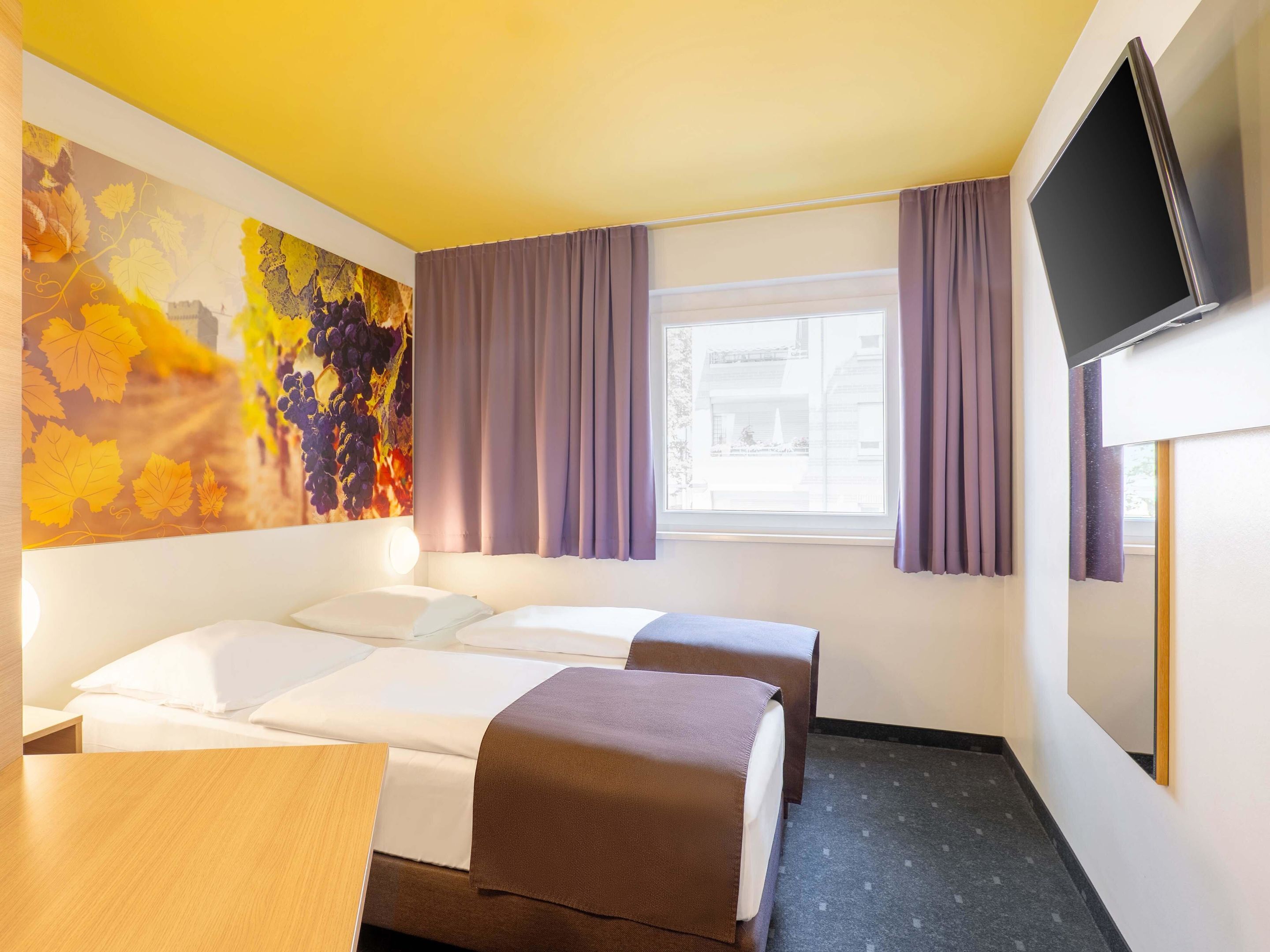 Hotellihuone, jossa on kaksi sänkyä, televisio, ikkuna, jossa on verhot, ja taulu seinällä.