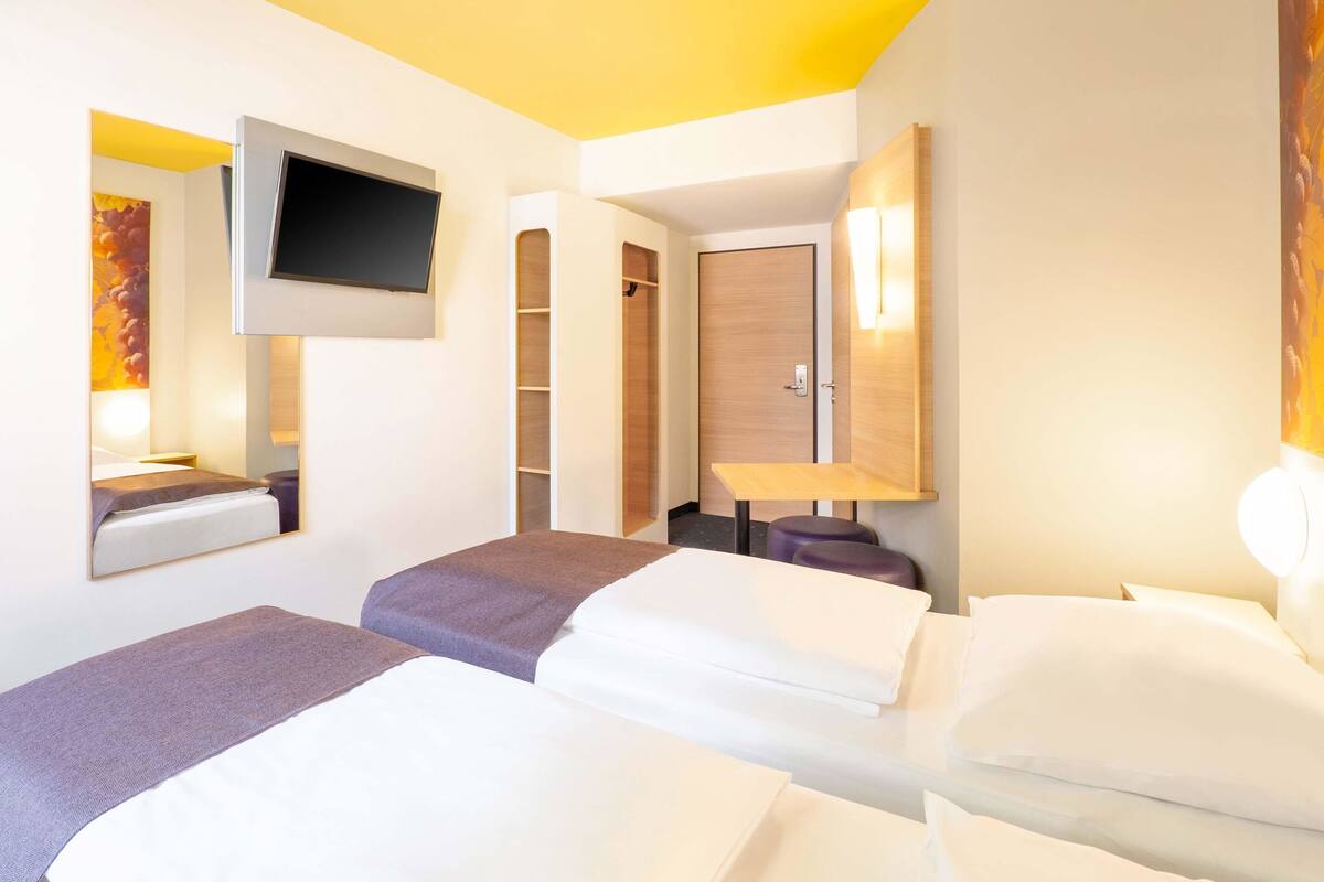 Quarto de hotel com duas camas, televisão presa na parede, guarda-roupa de madeira e uma mesa pequena.