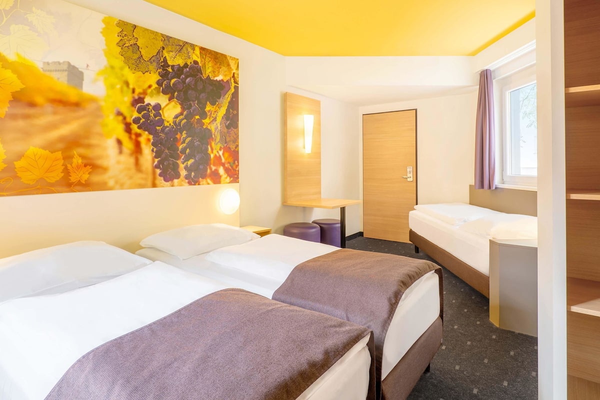 Quarto de hotel com duas camas, um quadro abstrato grande na parede, uma porta de madeira e uma janela com cortinas.