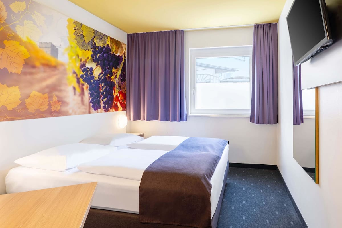 Quarto de hotel com uma cama, escrivaninha, televisão e janela com cortinas.