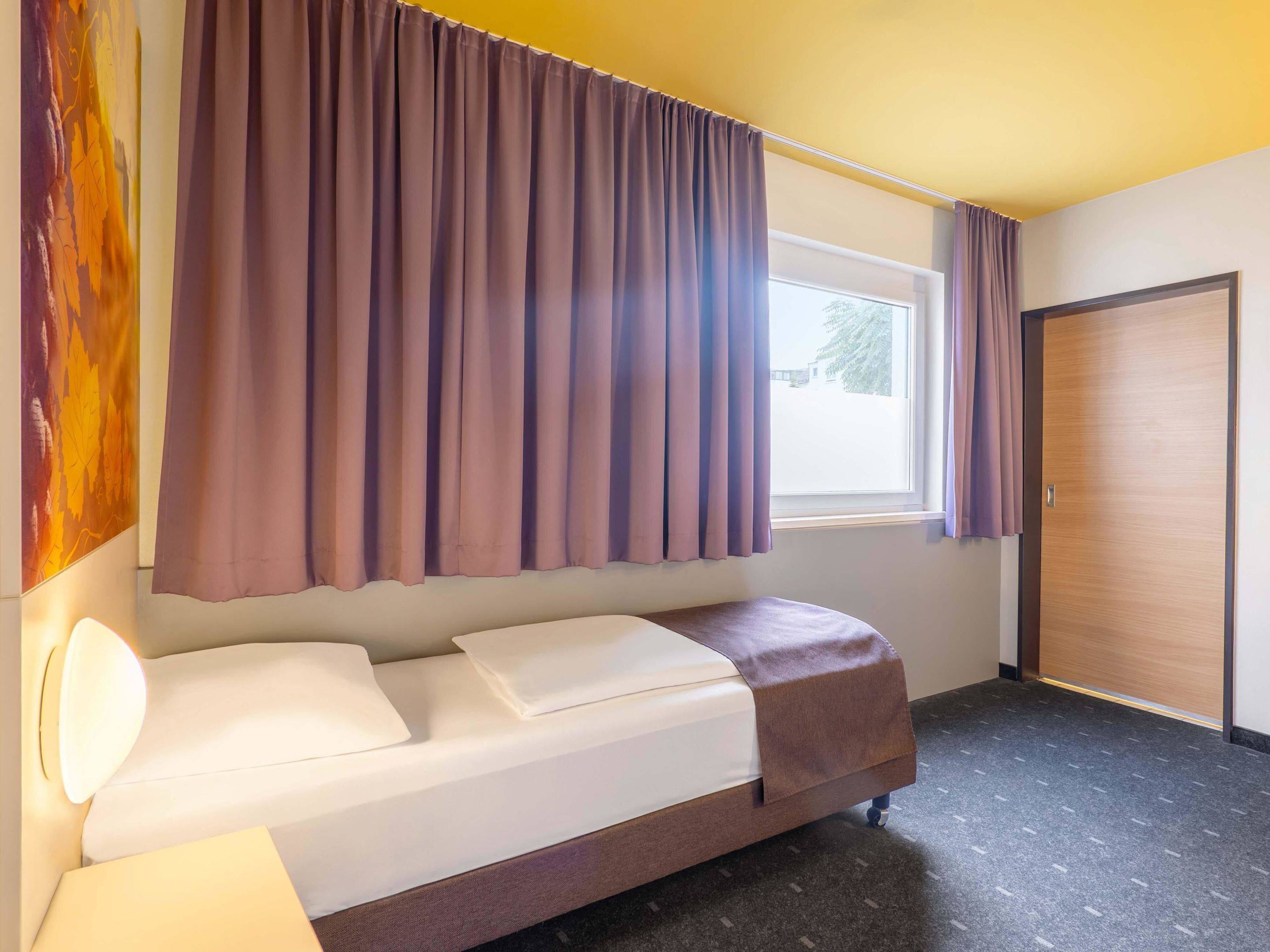 A single bed with a headboard, a window with curtains, a door, and a wall with a partial artwork.