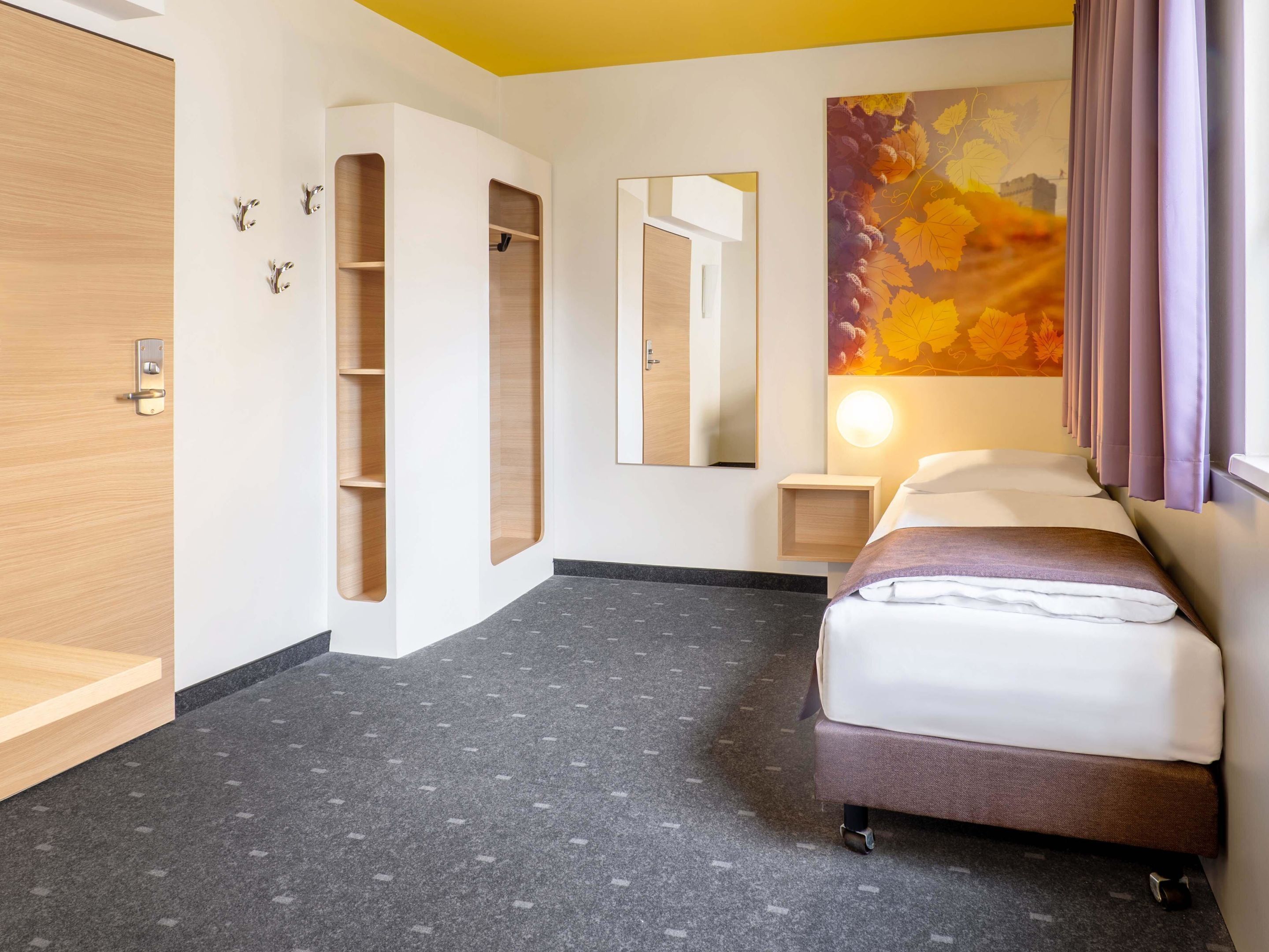 Quarto de hotel com cama, guarda-roupa, espelho, mesa de cabeceira e uma parede com arte decorada com padrão de folhas.