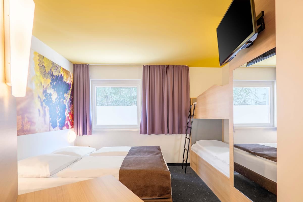 Quarto de hotel com beliche, cama de solteiro, escrivaninha e televisão fixada na parede.