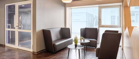 A modern lounge area with a sofa, two armchairs, and a small table with potted plants. There are large windows providing a view of the cityscape.