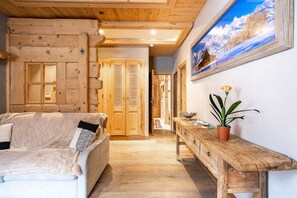 Ein gemütliches Zimmer mit Holzboden, einem Sofa mit Felldecke, einer Holzbank mit Topfpflanze und einem Gemälde einer verschneiten Berglandschaft.