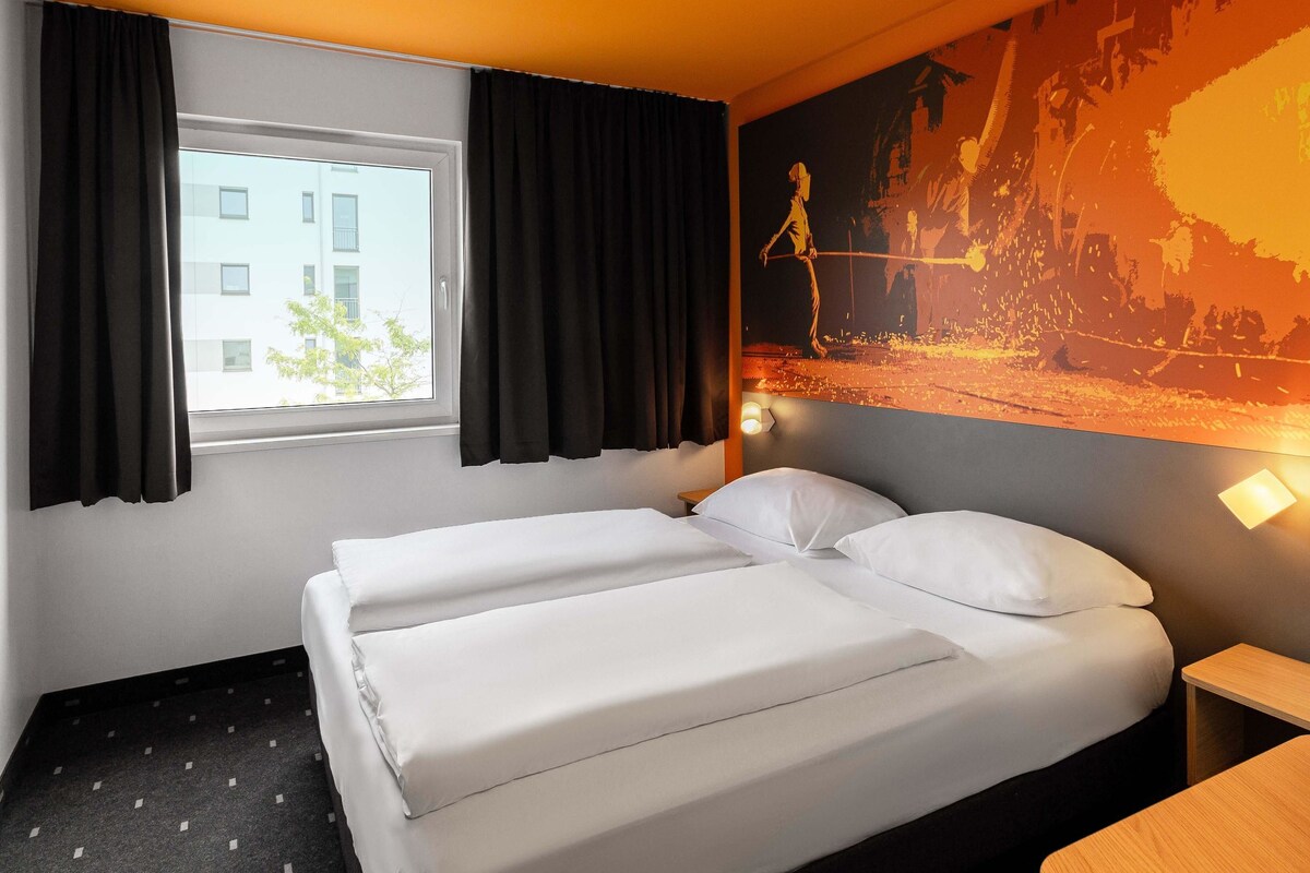 A hotel room with a bed, a nightstand, a window with curtains, and a wall mural.