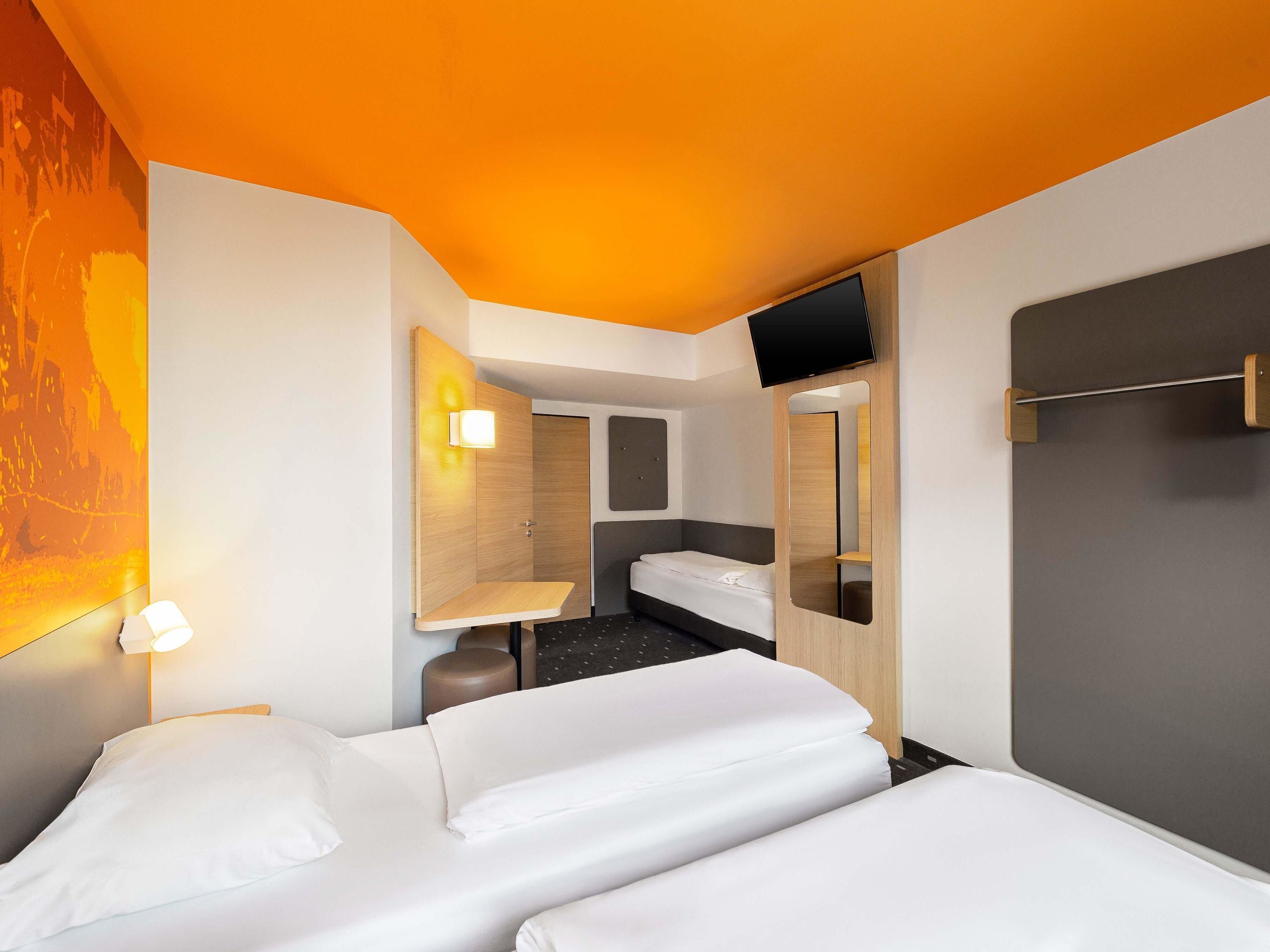 A hotel room with two beds, a bedside table, a mirror, a wall-mounted TV, and a small bathroom area.