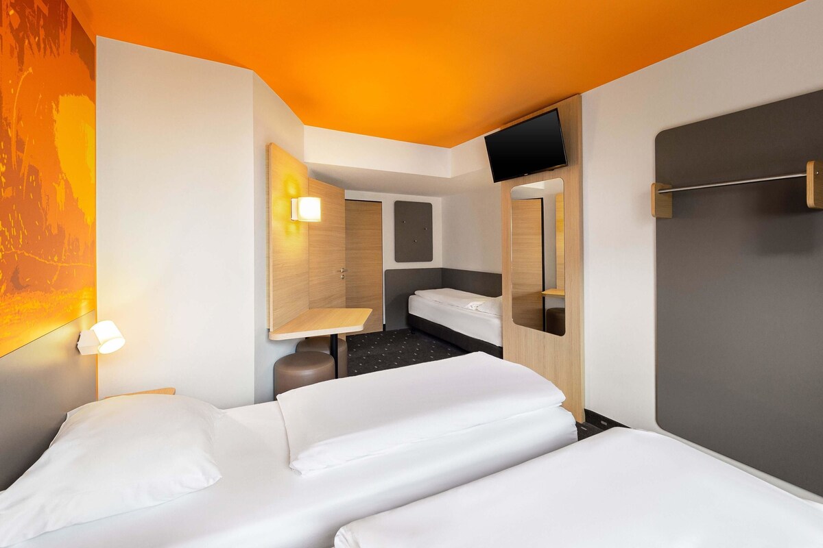 A hotel room with two beds, a bedside table, a mirror, a wall-mounted TV, and a small bathroom area.