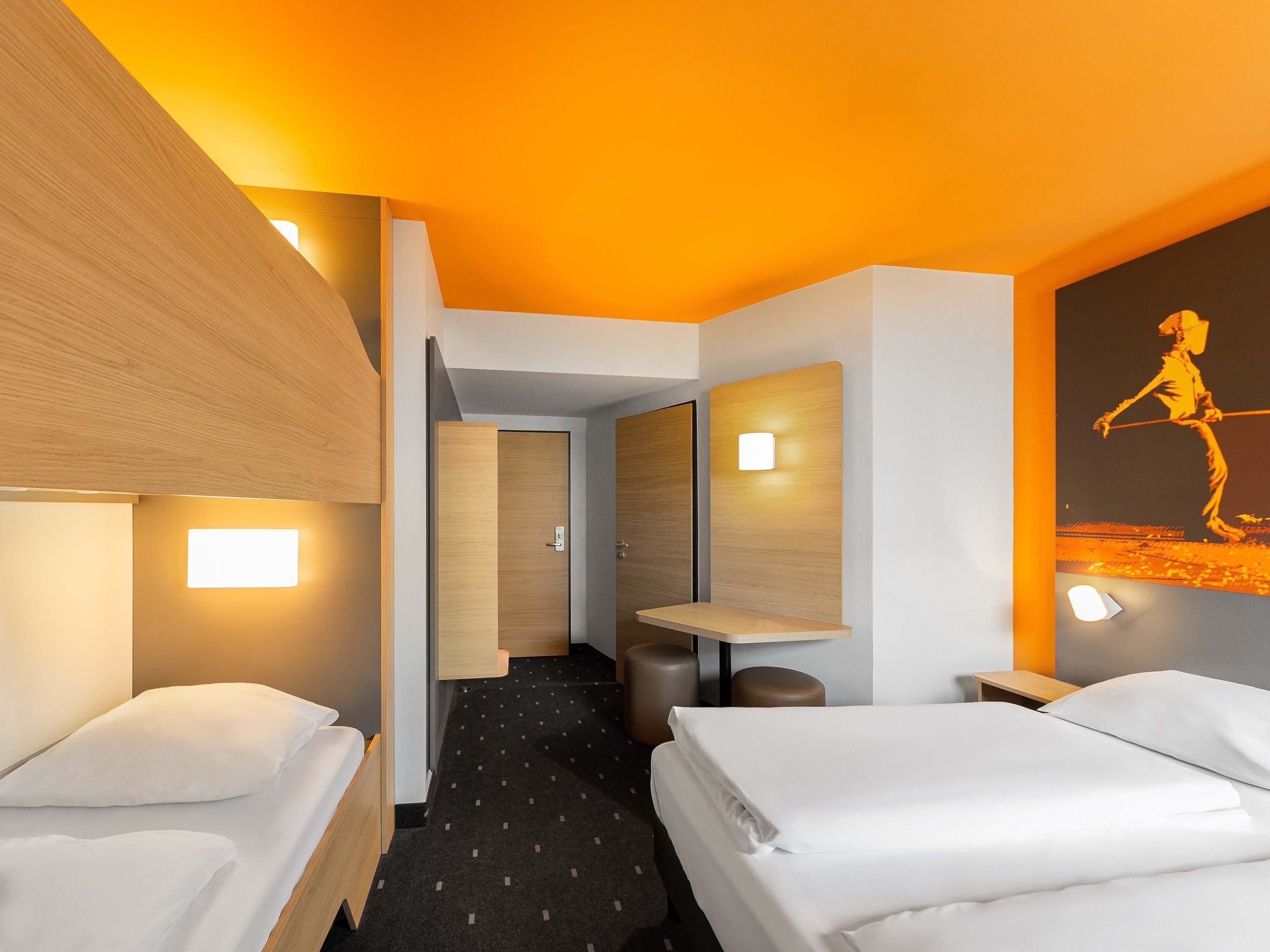Habitación de hotel con dos camas, un escritorio pequeño, una silla y una mesita de noche. Se ve un pasillo que conduce a otra habitación.