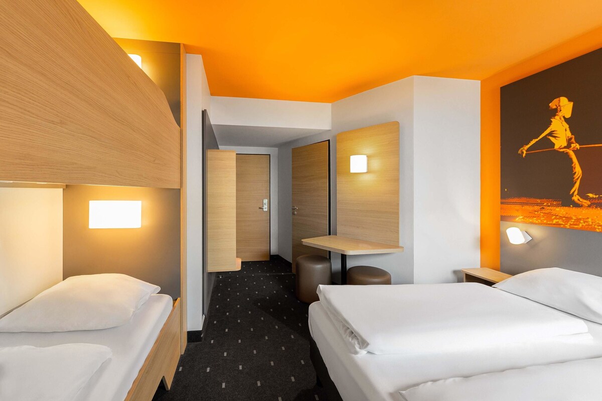 A hotel room with two beds, a small desk, a chair, and a nightstand. There is a hallway leading to another room.