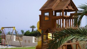 A wooden playhouse with a slide, a swing set, and a palm tree in the background. - Apartment for Five, Swimming Pool, Near the Beach (Rethymno)