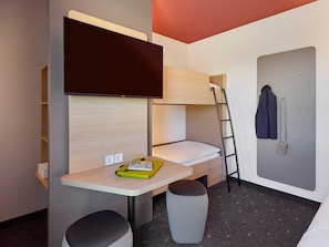 Una habitación de hotel compacta con una cama litera, un escritorio con silla, un taburete pequeño, un televisor instalado en la pared y un perchero.