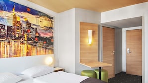 A hotel room with a bed, a nightstand, a wall-mounted light, a wooden shelf, a door, and a cityscape painting. - B&B Hotel Frankfurt City-Ost (Frankfurt)