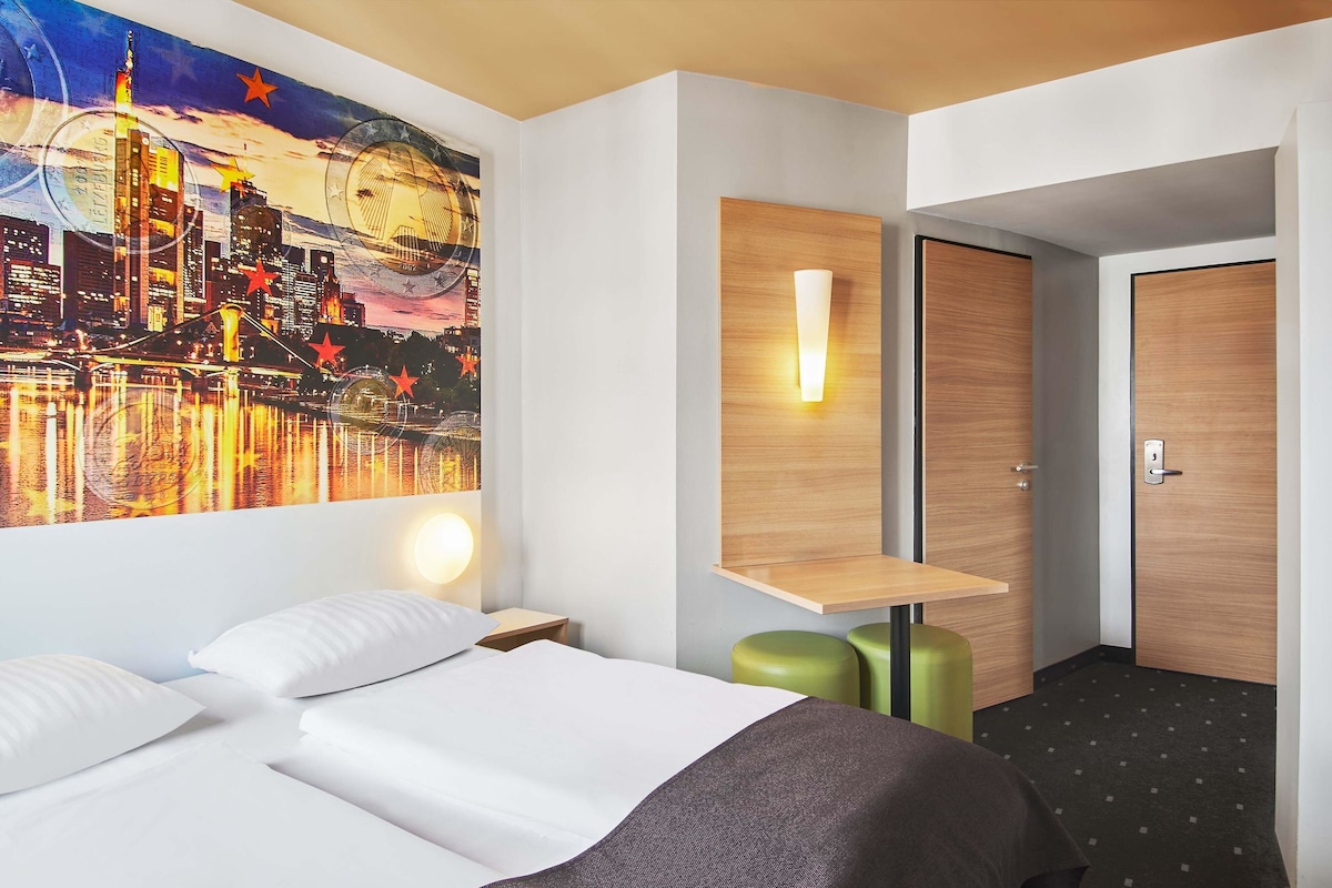 A hotel room with a bed, a nightstand, a wall-mounted light, a wooden shelf, a door, and a cityscape painting.