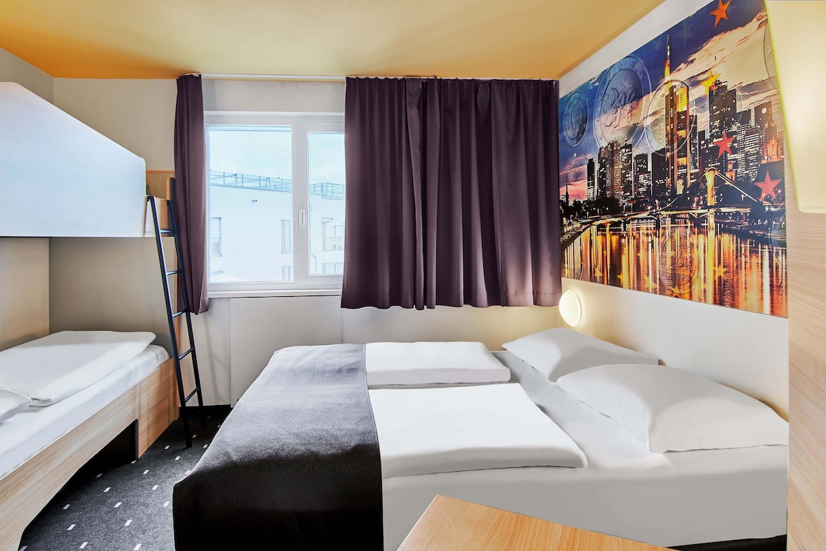 A hotel room with two single beds, a bunk bed, a desk, and a large window with curtains.