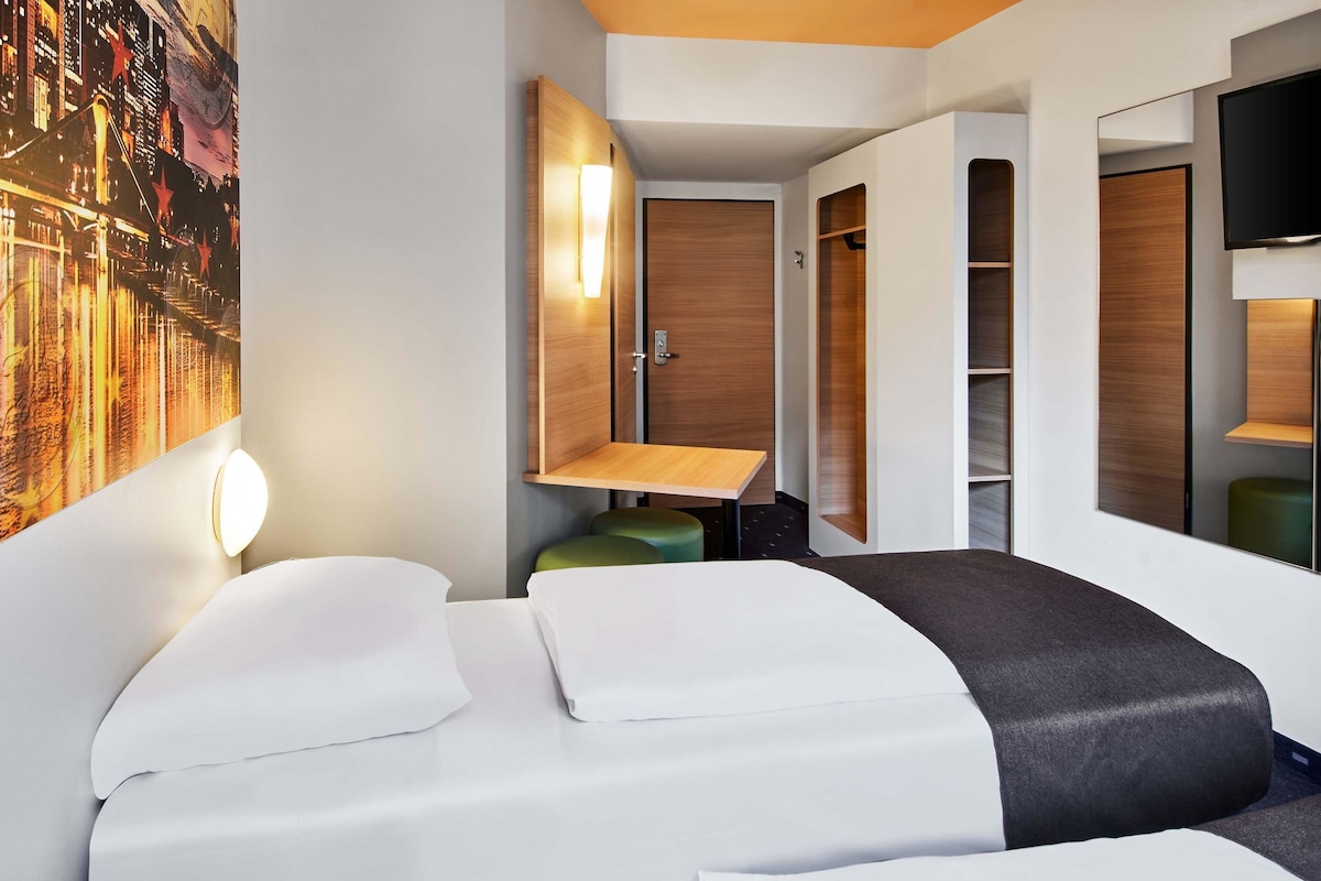 A modern hotel room with a bed, a bedside table, a mirror, a wall-mounted TV, and a large window.