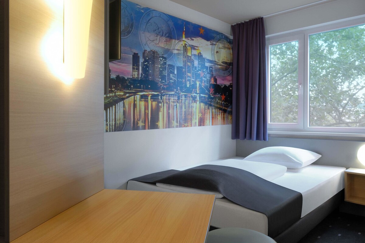 A hotel room with a bed, a desk, a window with curtains, and a cityscape mural on the wall.