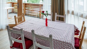 A dining area with a table set for four, wooden chairs, a red rose in a vase, and a view of a balcony with a wooden railing. - 1: Three-room Apartment With Panoramic View (Badia)