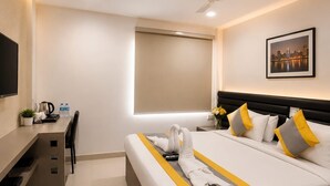 Quarto de hotel com cama, escrivaninha, cadeira, ventilador de teto, janela com persiana e um quadro na parede.