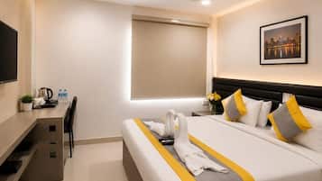A hotel room with a bed, a desk, a chair, a ceiling fan, a window with a blind, and a framed picture on the wall.