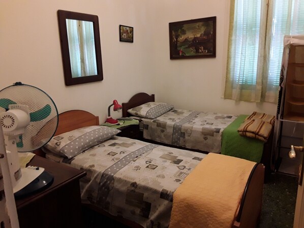 A hotel room with two single beds, a desk with a chair, a standing fan, a desk lamp, and a small painting on the wall.