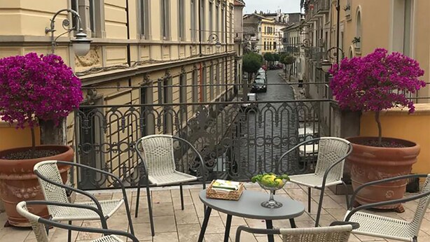 An outdoor terrace with wicker chairs and a table, surrounded by potted plants and a view of a narrow street.